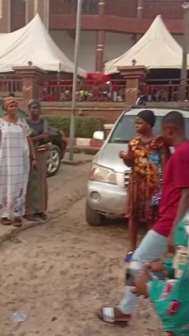 The video shows a gathering of people in what appears to be a public space. Several individuals are visible, including women dressed in colorful clothing, a boy in an orange shirt, and men in casual attire.  A large white tent with red and white decorations is set up in the background, possibly indicating an event. There are also cars and a motorcycle parked nearby. A building under construction is visible in the distance, and a banner with faces on it hangs near a building with a balcony.  The overall atmosphere seems lively and possibly celebratory.