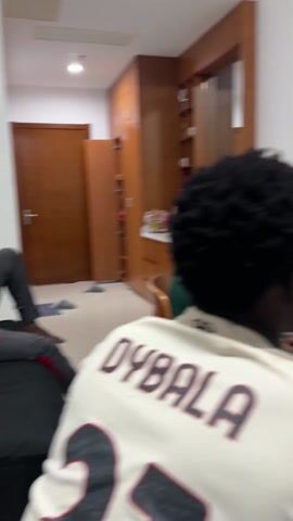 A man is sitting and playing a video game on a television in a room. The man is wearing a light colored shirt with the word "OYBALA" on the back, a green sleeve on his left arm and brown shorts. He has short, curly black hair. He is holding a game controller. The television is on top of a wooden dresser. A small refrigerator is in the background. The room is white and has wood trim.