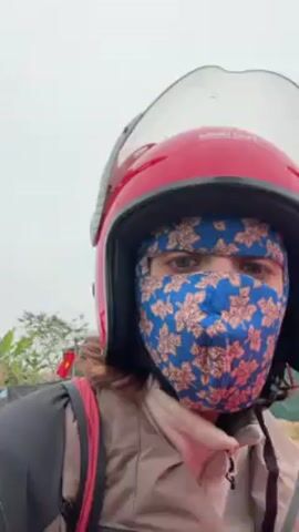 The video shows a person riding a moped on a road. The person is wearing a red helmet and a blue patterned face covering. The person has green eyes, and they are looking directly at the camera. The road is paved, and there are buildings and trees in the background. The sky is overcast.