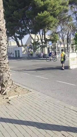 A group of cyclists are riding in a race on a paved road. The scene is outdoors, with trees and buildings in the background. The cyclists are wearing athletic clothing, and some are wearing helmets. The weather appears to be sunny and warm. The video shows multiple cyclists in motion, with one cyclist in the foreground wearing a yellow and black cycling outfit and a helmet, riding a dark-colored bicycle. Another cyclist in a white tank top and black shorts, also wearing a helmet, is riding a dark bicycle. In the background, several other cyclists are visible. A person wearing a bright yellow vest and dark shorts stands on the right side of the road, facing away from the camera, appearing to be a race official or volunteer. The road has painted lines and a pedestrian walkway made of interlocking bricks borders the road on the left. Traffic signs are visible in the distance, including a parking sign.