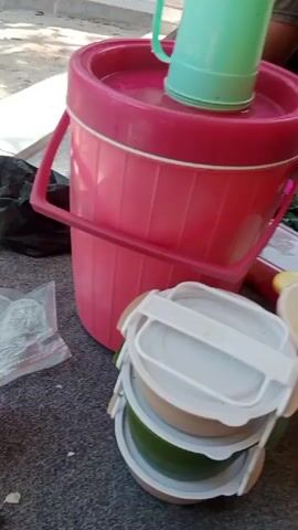 The video pans across a ground-level view of items arranged on a grey, carpeted surface outdoors. In the foreground, a stack of three food containers with white lids is visible. To the right of the food containers is a white bowl containing what appears to be food scraps, with yellow handles on either side. Further to the right, a blue and white rice cooker is plugged into an electrical outlet. Behind the rice cooker, a portion of a doorframe and wall is visible. To the left of the food containers, a large pink cylindrical container with a pink lid and a white rim stands upright. A green plastic cup rests on top of the pink container's lid. A pink bowl is visible near the top of the pink container. A small plastic bag containing white items is to the left of the pink container. A black trash bag is also present near the pink container. In the background, a person is partially visible, seated on the ground. Another person's leg is also visible near the blue rice cooker. The environment appears to be outdoors, possibly near a building entrance. The lighting suggests it is daytime.