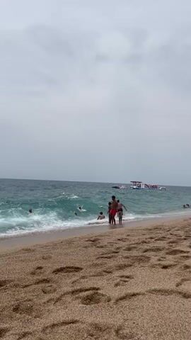 A wide shot captures a sandy beach with the ocean in the background. Several people are visible in the water, some swimming and others standing near the shore. Waves are crashing on the beach, creating a foamy white line along the water's edge. In the distance, a large inflatable water park can be seen in the ocean. The sky is overcast with clouds. A family of four, a man, woman and two children are walking along the shore towards the water. The man is wearing red swim trunks and the children are also wearing swim attire. They appear to be heading into the water for a swim.