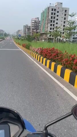 The video is filmed from the perspective of someone riding a blue motorcycle on a paved road. The road has a white line marking the lane and a curb with yellow and black stripes. Along the right side of the road, there is a grassy median with flowering bushes and several under-construction buildings in the background. A blue sign with a white "U" turn symbol and text indicating "KEEP RIGHT FOR U TOWN" is visible on the left side of the road. There are some orange traffic cones placed along the road ahead. The sky is overcast.