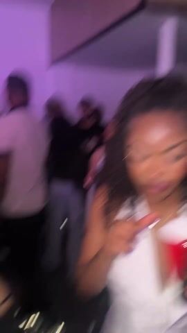 The video shows a woman dancing at a party. The scene is indoors with a purple-toned lighting. The woman is wearing a white halter top and has curly hair. She is smiling and raising her arms. Other people are visible in the background, some holding red cups. The camera moves, focusing on the woman's face as she dances.