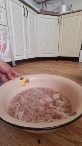 A close-up shot shows a person's hands interacting with a large, light-colored bowl filled with water and a substantial amount of thin, light-pink, tubular material, resembling intestines. The person is holding some of this material, which is being pulled out of the water and appears to be a continuous strand. The bowl is placed on a wooden floor. In the background, kitchen cabinets with white doors and a countertop are visible, suggesting the scene takes place in a kitchen. The lighting is bright.