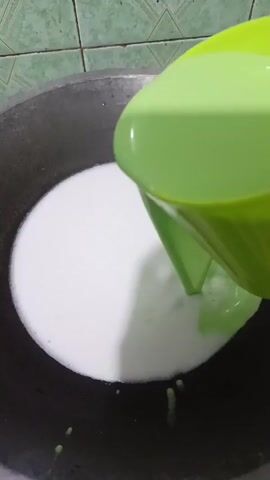 In this close-up, overhead shot, a light green liquid is being poured from a bright green pitcher into a dark gray pot. The pot contains a white liquid that is swirling and mixing with the green liquid, creating marbled patterns. The liquids appear to be of a similar consistency, possibly a dairy-based mixture or a dessert ingredient.

The scene is likely taking place in a kitchen, suggested by the tiled background with green and white squares, a common feature in Indonesian kitchens. The lighting appears to be natural, casting subtle shadows on the liquids, indicating it might be daytime.

The activity captured is the preparation of food or a beverage, where two liquids are being combined. The focus is on the visual interaction of the liquids as they merge, highlighting the textures and the creation of swirls. There are no people visible in the frame, and no text is apparent. The overall impression is one of culinary creation and simple domestic activity in a typical Indonesian setting.