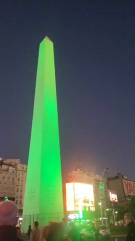 Here's a description of the video:

The video shows the Obelisco de Buenos Aires at night. It is lit up in a vibrant green color. People can be seen walking around the base of the obelisk. In the background, there are buildings and a large digital screen also lit up. The sky is a dark shade of blue. A few cars can be seen driving in the street.
