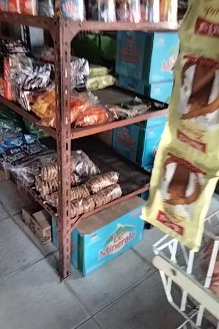 The video shows a close-up view of a store shelf displaying various packaged goods, primarily snacks and instant noodles. The shelf is made of rusty metal and is divided into compartments. On the top shelf, there are several blue boxes with images of food. Below that, there are rows of packaged snacks, some in clear plastic and others in colorful wrappers. There are also stacks of cookies and bags of chips. The lighting is dim, and the camera is in motion, suggesting it is handheld. The overall impression is of a small, cluttered shop.