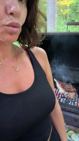 The video shows a close-up of a woman with dark hair and light skin wearing a black tank top and a gold necklace. She is standing in front of an outdoor grill with a steak cooking on it. The steak is thick and appears to be a ribeye cut, with visible grill marks. Smoke is rising from the grill. The background is blurred but suggests a wooded outdoor setting. The woman is looking towards the camera and licking her lips, with a slight smile. The overall impression is that she is about to eat or enjoy the steak.