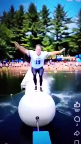 This media file captures a group of people engaged in recreational activities at an outdoor water park. The scene is set on a bright, sunny day, with lush green trees forming a backdrop. The water appears calm and clear, reflecting the blue sky.

The main subjects are individuals participating in various challenges. One person is seen on an inflatable floating platform, appearing to balance with arms outstretched. Another individual is on a blue beam suspended over the water, also attempting to maintain balance. A third person is shown swinging from a rope attached to a large hoop, demonstrating athleticism. The overall mood seems to be one of fun and adventure, with participants likely experiencing excitement and perhaps a bit of thrill.

The activity appears to be a form of obstacle course or water-based challenge designed for recreation and entertainment. The presence of other people on a dock-like structure in the background suggests this is a public or communal space for such activities.

Visible text includes the number "19" on the shirt of one participant and "9" on the shirt of another. There are also icons resembling a "like" button with the number "107" next to it, a speech bubble with "2" next to it, and a share icon, indicating the media may be from a social media platform. While the context mentions Takum, Nigeria, there are no specific visual cues in the video that definitively confirm this location. The setting, with its water, trees, and recreational structures, is consistent with many outdoor water parks found globally.