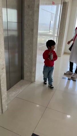 A young child stands in front of an elevator, holding a small, metallic container. The child wears a red sweater with a cartoon graphic, baggy blue jeans, and multi-colored sneakers. The elevator door is silver, and the surrounding wall has a speckled pattern. The floor is tiled. Another person stands slightly out of focus in the background to the right. The child looks directly at the camera, then down and to the side. A voice is heard speaking in the background, presumably addressing the child.
