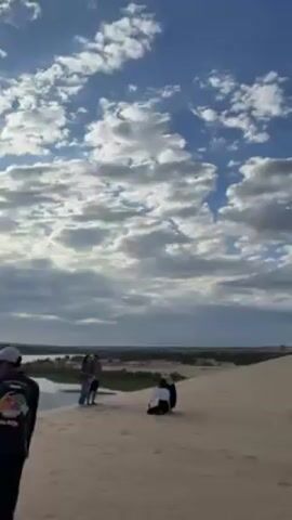 The video shows a sand dune landscape under a bright blue sky filled with fluffy white clouds. Initially, three people stand at the edge of the dune while another kneels, potentially taking a photo. The camera then pans to the right, revealing a yellow off-road vehicle parked on the sand. Other people can be seen scattered further up the dune. There are tire tracks in the sand, suggesting the vehicle has been driven on the dunes. The overall environment is arid and sandy, with some vegetation visible in the distance at the edge of the dunes.