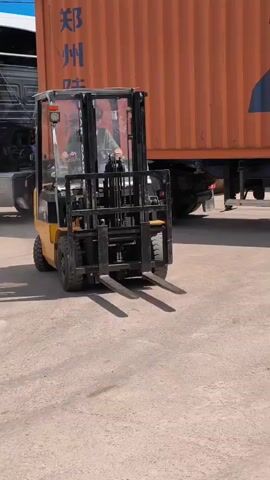 A man operates a yellow and black forklift (model CPCD 15 CG, branded "CARGADOR") in an outdoor industrial yard, likely in Molokovo, Russia. The forklift moves a short distance forward, with its forks lowered, in front of a large orange shipping container. The container is partially visible, showing some Chinese characters (郑州, indicating Zhengzhou) and English text including a telephone number prefix and a web address (WWW.ZZGUO). The scene takes place during the daytime under clear, sunny weather conditions, suggesting a busy loading or unloading area. The forklift operator, dressed in a green shirt and dark pants, appears focused on the task. A portion of a truck's wheel is visible behind the container, suggesting the container is mounted on a trailer.