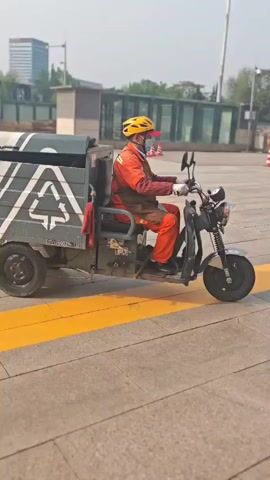 A three-wheeled electric cargo vehicle is driven by a person wearing an orange uniform, a yellow safety helmet, a blue face mask, and gloves. The vehicle has a grey and white cargo box on the back, featuring a prominent recycling symbol on the side. Chinese characters "环卫作业 随时停靠" (Environmental Sanitation Operation, park anytime) and "京A2000HZ-332" are visible on the rear of the box.

The video, filmed during daytime with bright sunlight, shows the vehicle first moving from right to left across a paved area, then turning away from the camera and proceeding further into the background. In the background, a blue sign with "Taxi" in English and "出租车候车区" (Taxi Waiting Area) in Chinese is visible, indicating a transportation hub or station. Behind the sign is a covered structure, likely a waiting area, and an ambulance can be seen parked. Other buildings and some trees are also visible further in the distance. The overall scene suggests an urban or semi-urban environment in Fengtai District, China, where environmental sanitation or recycling services are being carried out.
