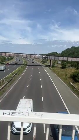 The video shows a highway with multiple lanes in both directions. Cars and vans are driving on the highway under a partly cloudy sky. There is a pedestrian bridge over the highway. The video appears to be shot from the pedestrian bridge, looking down at the highway. The highway is surrounded by greenery and trees. The video is short and does not show any people or specific interactions.