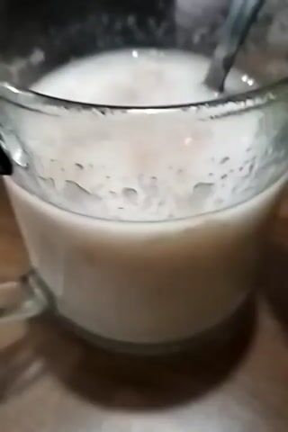 A close-up shot shows a clear glass mug filled with a light-colored liquid, possibly milk or a milky drink. A metal spoon is submerged in the liquid, and the liquid appears to be gently stirred. The mug rests on a wooden surface, likely a table. The background is blurred, but suggests an indoor setting with some objects and a light source. The overall impression is of someone preparing or consuming a beverage.