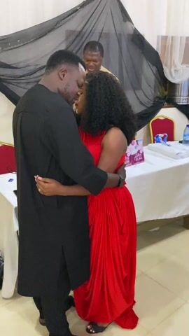 The video shows several couples dancing in what appears to be an event space. Initially, a man in a long black outfit is dancing with a woman in a red dress. They are close, embracing. The camera shifts to show another couple dancing; the woman is in a black dress, and the man is wearing a white shirt, vest, and grey pants. The room has a backdrop of black fabric and red and gold chairs. A table covered with a white tablecloth and some items on it is also visible.