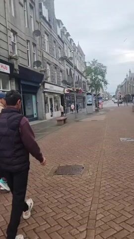The video shows a pedestrian walkway in a city center. The walkway is paved with red bricks laid in a herringbone pattern. The buildings lining the walkway are a mix of old and modern architecture. On the left, there are buildings with stone facades and traditional shopfronts. On the right, there is a large, modern building with a dark, textured facade, and further down, a shopping center with the name "BON ACCORD" displayed prominently. Several people are walking along the street. There are also streetlights, benches, flower baskets, and a bicycle rack with a bicycle. An advertising display is also visible. The sky is overcast, suggesting a cloudy day.
