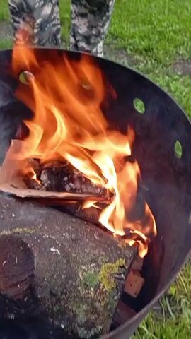 A fire burns in a black metal barrel. A log rests at the bottom of the barrel. A person wearing camouflage pants stands behind the barrel. The fire is mostly orange and yellow flames, with some smoke visible. The environment appears to be outdoors, with grass visible in the background.