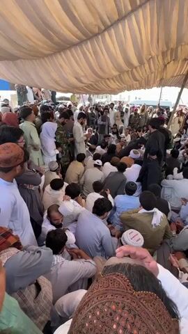 A large gathering of people, mostly men, are seated and standing under a beige tent. The crowd is densely packed, with many individuals wearing traditional clothing like shalwar kameez and kufis. In the center of the gathering, several individuals appear to be engaged in a discussion or meeting. Some are standing, including individuals in military-style uniforms, while others are seated on the ground. The environment is outdoors, suggested by the bright lighting and the tent structure. The camera perspective is from within the crowd, looking upwards towards the center of the assembly.