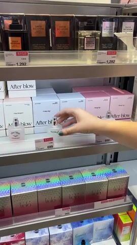 A person's hand is seen selecting a small, clear bottle of perfume from a shelf in a store. The shelf displays various perfumes in boxes, some with "SALE" tags. In the background, more perfume bottles and boxes are visible on other shelves. The overall environment suggests a perfume or beauty product retail setting.