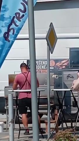 A man in a red t-shirt and black shorts stands in front of a grill, wearing an apron. A woman with gray hair sits at a table, looking towards the grill. A dog, possibly a German Shepherd, is visible behind the table. In the background, a food truck or stand is partially visible, with smoke emanating from it. A blue banner with white text is also visible on the left side of the frame. A diamond-shaped road sign is mounted on a pole in the center. The setting appears to be outdoors during the daytime.