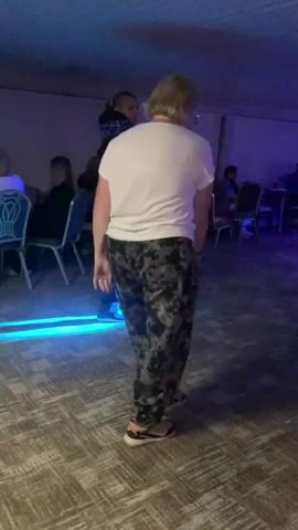 The video shows a group of people in a dimly lit room with blue and purple lighting. The room appears to be a dance floor or event space, with tables and chairs set up along the walls. Several people are dancing in the center of the room, while others are seated at tables. The individuals are of various ages and genders, dressed in casual attire. The lighting creates a party atmosphere.
