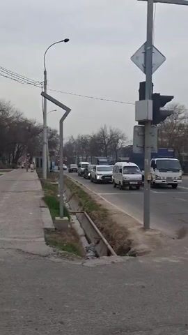 The video shows a traffic stop in what appears to be a city. The camera pans from a line of cars and a traffic light to the left.  The cars include a white van and a small truck. The camera then pans to the right to a crosswalk and a building that says “korzinka.uz.”  Several cars are parked in front of the building.  A few people are visible near the crosswalk.