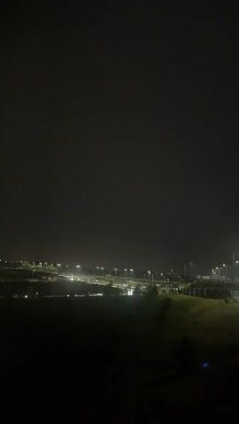 The video shows a dark night scene with a dimly lit landscape in the distance. Several rows of lights, possibly from a parking lot or industrial area, are visible, creating a hazy glow. Streetlights are also present, illuminating parts of the road and surrounding vegetation. The sky is completely dark, indicating a lack of moonlight or stars. The foreground is almost entirely black, with faint outlines of bushes or trees suggesting a natural landscape. The overall impression is one of an urban or suburban area at night, possibly with some rain or fog contributing to the diffused lighting.