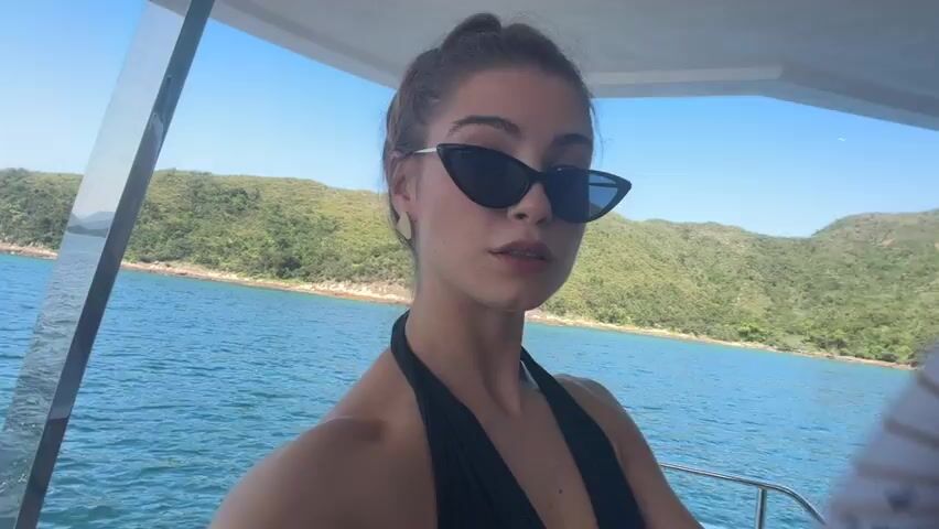 A young woman with dark hair pulled up, wearing sunglasses and a black swimsuit, is holding a white fan with a floral pattern. She is on a boat, with the ocean and lush green hills visible in the background under a clear blue sky. She is looking at the camera and fanning herself.