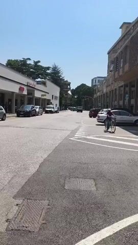 This video captures a street scene in Padua, Italy, on a clear, sunny day, likely late morning or early afternoon given the bright sunlight and distinct shadows. The camera pans across a moderately busy street lined with commercial buildings and parked cars.

The main subjects are the street, vehicles, and pedestrians. Several cars are visible, including a silver hatchback, a dark sedan, a blue hatchback, and a white sedan. A person is riding a bicycle across a crosswalk, wearing a white t-shirt and patterned shorts. Another person, wearing a beige cap and pants, is walking across the street, holding what appears to be a phone to their ear. There are also bicycles parked along the sidewalk.

The setting is a commercial area. On the left side of the street, there's a building with several storefronts, including one with a prominent green and yellow sign that reads "Prix Qualità Italiana". Another storefront displays an orange sign with Italian text that seems to indicate food businesses: "la Macelleria la Gastronomia la Panetteria". The architecture suggests a mix of modern and older buildings.

The activity is typical urban street life, with people going about their day, shopping, or commuting. The overall mood is relaxed and ordinary, reflecting a normal day in the city. The clear sky and bright sun suggest pleasant weather conditions.