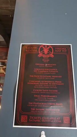 The video shows two flyers for a "Satanic Revival" event. The top flyer is for Friday, May 1st, and lists several events, including an opening ceremony, campaign updates, a path to ministry, an awards ceremony, music performances, and a drag performance. The bottom flyer is for Saturday, May 2nd, and includes events such as a discussion on transhumanism, testimonies, a speech about Satanic religion, a Q&A with the Executive Ministry, and several musical performances. Both flyers feature the Satanic Temple logo and advertise ticket availability at "satanic-revival.com". They are taped to a wall with a small white hook in between. The camera zooms in slightly to focus on the flyers. Sounds of a crowded and noisy room can be heard.