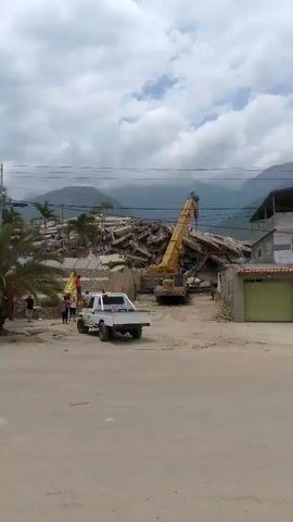 A white pickup truck is parked on a dirt road in front of a large pile of rubble. A yellow crane is positioned in front of the rubble, with its boom extended over the debris. Several people are visible in the background, standing near the rubble. There are palm trees and other vegetation in the foreground and mid-ground. Mountains are visible in the background, and the sky is cloudy. The overall scene suggests a construction or demolition site, or possibly the aftermath of a disaster.