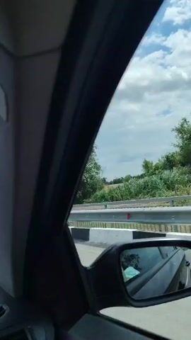 The video shows a first-person perspective from inside a car driving on a highway. The driver's hands are on the steering wheel, and the dashboard with gauges is visible. In the rearview mirror, a person's face is reflected, holding a red object, possibly a phone. Outside the car, a cloudy sky and a rural landscape with fields and trees can be seen. A yellow car is visible on the road ahead. Various car accessories, such as an air freshener and a compass, hang from the rearview mirror and the dashboard. The scene suggests a road trip or a drive through the countryside.