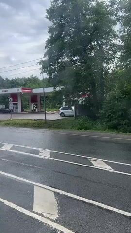A black hatchback vehicle is seen from a side view driving on a wet asphalt road. The road appears to have tram tracks. In the background, there is a gas station with red and white branding. To the right of the gas station, there are trees and a white vehicle. Further down the road, a bus stop shelter with a person sitting inside is visible, also surrounded by trees. The weather appears overcast and rainy, with water visible on the road surface.
