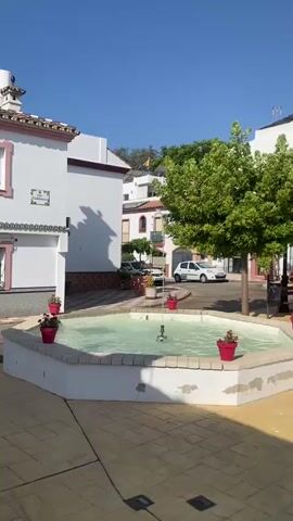 The video shows a street scene with a fountain in the foreground. Water shoots up from the fountain, and the spray is visible. There are potted plants around the fountain, some with red flowers. Behind the fountain, there is a sidewalk with a patterned tile design. Two cars are parked on the street, a white SUV and a white hatchback. A large green tree is on the right side of the frame, and a white building with red trim is on the left. In the background, there are more buildings, some with balconies, and a Spanish flag is flying from a rooftop. The sky is blue and clear. The scene appears to be a sunny day in a town or village.
