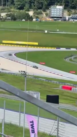 The video captures a motorsport race from an elevated spectator viewpoint. Two race cars are visible on a track that winds across a grassy landscape. The foreground features sections of a metal railing and a pink flag bearing the logo “BWL.” Beyond the railing, the track is bordered by red and white stripes and is set against a backdrop of green hills and a structure with a building on it, on the background on the right. The camera appears to be steady, providing a stable view of the racing action as the cars navigate the course. A person can be seen at the bottom left of the screen, partially obstructing the view of the track.