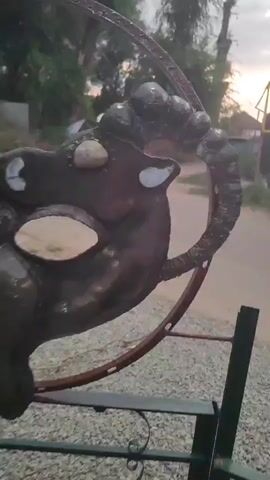 A close up panning shot of a dark metal sculpture of a mask on a rusty frame outside. The mask appears to be a stylized face with large eye openings and bumps on its forehead. The sculpture is set against a background of gravel, trees, and a fence with a house beyond it. The sky is visible and shows signs of dusk.