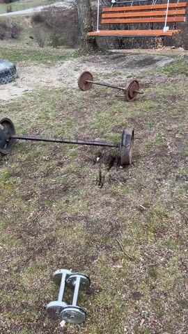 The video shows an outdoor weightlifting setup.  It starts with a view of two barbells with weights lying on a grassy area, with a wooden swing set in the background. The camera then pans over to a weight bench with a barbell and weights on it, followed by a table with dumbbells and weights under a structure with a fence. Finally, the camera shows a gray car parked on a grassy field, with a line of trees on a hill in the background under a cloudy sky. There are no people visible in the video.
