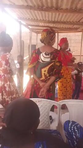 The video shows a group of people gathered outdoors, under a corrugated metal roof. There are four women standing: one wearing a red and yellow patterned dress with a red headwrap and speaking to the others, another wearing a red and brown patterned dress and carrying a baby on her back in a carrier, one in a brown and red floral dress, and one wearing a red dress and yellow patterned skirt while holding a baby. There are two children standing near the woman in red and yellow, dressed in casual clothing. Several white plastic chairs are visible in the foreground, with an individual in a blue and white dress seated in one of them. The setting appears to be a backyard or outdoor gathering space, with a brick wall in the background. The woman in red and yellow appears to be giving a speech or presentation.