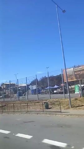 The video, shot from a moving vehicle, captures a landscape marked by destruction and aftermath. Initially, the view shows a guarded area with fencing, temporary structures, and various vehicles. The scene then shifts to the charred remains of a hillside, with burnt trees and debris.  The destruction indicates a fire or other devastating event.  Throughout the video, the sky is a clear blue.