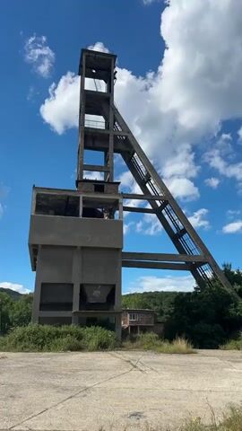 A concrete mining headframe stands against a blue sky with fluffy white clouds. The structure is tall and angular, with a narrow, open-air tower at the top supported by a sloped truss. Below the tower is a rectangular, multi-story concrete building, also open and showing signs of decay. Overgrown vegetation surrounds the base of the structure and the surrounding paved area. In the background, a tree-covered hill and a small, brick building are visible. The scene appears to be in an abandoned or disused industrial area. There are no people visible in the video.