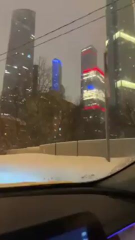 A video shot from inside a vehicle shows a snowy city landscape. Tall, modern skyscrapers dominate the view, some illuminated with blue and red lights. Snow is visible on the road and the car's windshield. The car's dashboard and infotainment screen are visible, displaying the name of a song titled "Vrana". The car is moving forward on the snow-covered road.