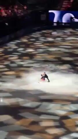 The video shows two ice skaters on an ice rink during what appears to be a performance. The ice rink has a patterned projection on it, with what looks like the Olympic rings at one point. The skaters are wearing performance costumes: a dark suit for one and a red and dark dress for the other. They move in unison, engaging in maneuvers that suggest a pairs routine. The setting suggests a large indoor arena or stadium, with an audience visible in the background, adding to the atmosphere of a formal, public event.