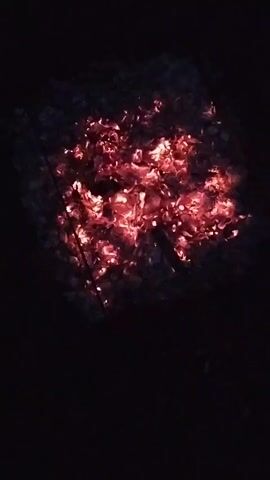 The video shows a top-down view of glowing red embers in darkness. The embers are clustered together, forming a rough square shape. There are hints of ash and charcoal surrounding the glowing areas. The lighting is dim, with the light from the embers being the primary source of illumination.