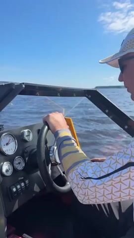 The video shows a man operating a boat with another person in the passenger seat. The man steering the boat is wearing sunglasses and a white baseball cap with gold trim. He is also wearing a white long-sleeved shirt with a gold star pattern. The boat is moving across a body of water, with the wake visible behind it. The sky is blue with some clouds. In the background, there is a shoreline with trees. The passenger in the boat is a woman wearing a white baseball cap and a black long-sleeved shirt with a gold and white pattern. She appears to be holding something small in her hands. Another person, a man, is sitting behind the woman, also wearing sunglasses and a black long-sleeved shirt with a gold and white pattern. There are fishing rods visible in the boat.