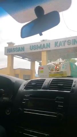 The video shows a car driving towards the entrance of Usman Katsina International Airport. The view is from inside the car, looking out through the windshield. The airport entrance has a sign that reads "USMAN KATSINA INT'L AIRPORT" in green lettering. There is a box of tissues with a golden deer decoration on the dashboard of the car. The weather appears to be overcast.