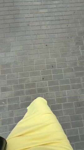The video shows a person walking on a paved sidewalk. The camera is positioned looking down at the person's feet and lower legs. The person is wearing a long, bright yellow dress and black sandals. A black purse is visible on the left side of the frame, being held by the person. The sidewalk is made of rectangular gray bricks laid in a stretcher bond pattern. The lighting suggests it is daytime. The action is limited to the person's feet and legs moving forward, indicating walking. There are no other people or objects clearly visible in the frame.