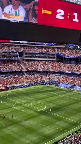 This is a wide-angle, high-angle shot of a soccer stadium during a match. In the foreground, a vibrant green soccer field is visible with a few players scattered across it. The field is marked with white lines, including a center circle and penalty boxes. Above the field, a large red scoreboard displays the score "2:1" with flag icons representing Spain and Belgium. The scoreboard also shows images of a couple, identified as "Jordan Love" and "Hisense". The stadium is packed with spectators, mostly wearing red, filling the tiered seating that surrounds the field. The atmosphere appears lively, suggesting a significant event is taking place. The lighting suggests it is daytime, possibly under an open or partially covered stadium roof.
