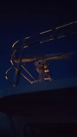 The video shows the radar system of a boat at night, rotating against a dark blue sky. The radar antenna, a long, thin bar, spins continuously atop a white pedestal on the boat's roof.  A metal railing illuminated with a warm light surrounds the radar apparatus. The rest of the boat is shrouded in darkness, visible only as a black mass beneath the radar installation.